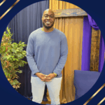 Black man with glasses, long sleeve blue shirt, and light blue pants standing in front of dark blue and gold wall of fabric