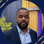 Black man with navy blue jacket and light blue collared shirt standing in front of dark blue and gold wall of fabric