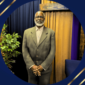 Black man with glasses, grey beard, olive colored suit, light colored shirt standing in front of a dark blue and gold curtained wall