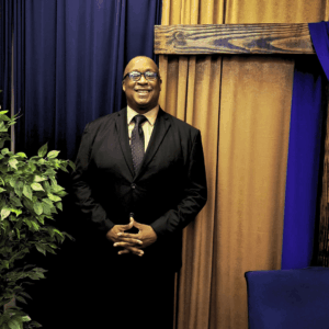 Black Man With Black Suit, Light Brown Shirt and Black Tie wearing glasses and standing against a dark blue and gold curtained wall