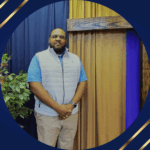 Black man with Blue shirt and lighter blue zipper vest standing in front of a dark blue and gold curtained wall
