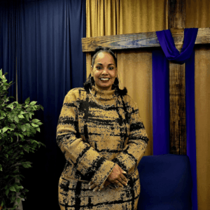 Black woman with gold and black blouse, dark curled hair, and gold and black skirt standing against a dark blue and gold curtained wall