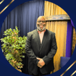 Black man with black suit and salmon colored shirt standing in front of dark blue and gold curtained wall