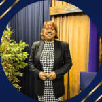 Black woman with short auburn hair in a black dress with a black and white dress standing in front of a dark blue and gold curtained wall