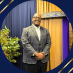 Black man with glasses, grey plaid jacket, white collared shirt, and black suit pants standing in front of dark blue and gold curtained wall