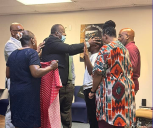 Black Bishop in Black Suit laying hands on church member in white shirt and black pants surrounded by members and ministers