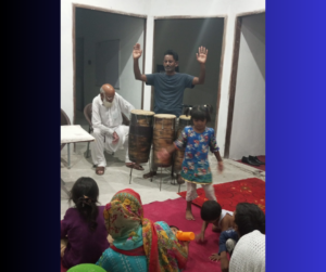 Older Pakistani man in all white, younger Pakistani man in dark green shirt playing instrument and worshipping God
