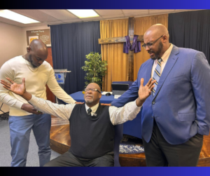 Three black men, two standing in blue suit jacket, other in beige shirt with jeans laying hands on a seated man with black vest and lightly colored shirt