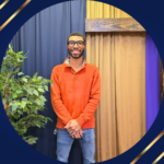 Black man in orange sweater and blue jeans standing in front of a dark blue and gold curtained wall
