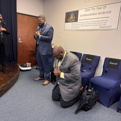 Black minister kneeling in pray in gray and black suit, black minister in dark blue suit bowing head in prayer